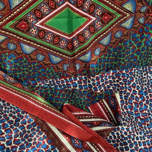 Może przedstawiać: Zbliżenie kolorowej tkaniny z geometrycznym wzorem rombu w kolorze zielonym, czerwonym i białym. Tkanina posiada również wzór w panterkę w kolorze niebieskim i czerwonym. Widoczna jest również czerwona wstążka z białymi i brązowymi paskami.