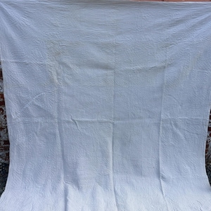 May include: A large, rectangular white quilt featuring an all-over quilted texture with visible stitching patterns. The fabric has a slightly wrinkled appearance and is displayed hanging vertically, showing a uniform solid white color throughout.