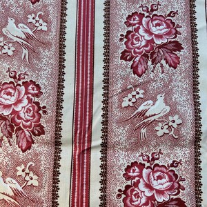 Op de afbeelding: Een crèmekleurige stof met een herhalend patroon van rode rozen, vogels en bloemmotieven. Verticale strepen in rood en bruin dragen bij aan het vintage ontwerp. De stof heeft een gestippelde textuur, wat een klassieke uitstraling creëert.