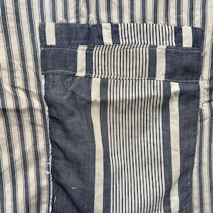 Peut inclure: Gros plan sur un tissu à rayures verticales bleu marine et blanc cassé. Une poche bleu marine est cousue sur le tissu, avec un motif rayé similaire. Le tissu semble légèrement usé, avec quelques effilochages sur les bords.