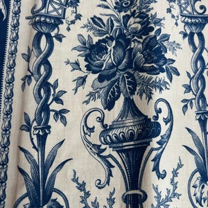 Könnte beinhalten: Nahaufnahme eines Stoffes mit blau-weißem floralem Toile-Muster. Das Design zeigt eine große Vase, die mit Rosen überquillt, umgeben von architektonischen Elementen und Ranken. Der Stoff hat eine leinenartige Textur.