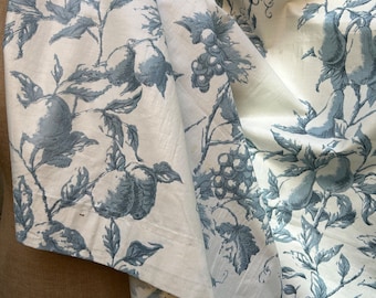 May include: Off-white fabric with a blue fruit and foliage pattern. The design features pears, grapes, and leaves. The fabric is draped, showing the detailed print and texture. The overall theme is a classic, botanical design.
