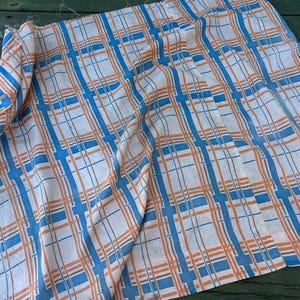 May include: A white fabric featuring a plaid design in blue and orange. The fabric is draped, revealing a pattern of intersecting lines. The blue lines are broader than the orange ones, forming a grid-like pattern. The fabric appears to be lightweight and suitable for clothing.