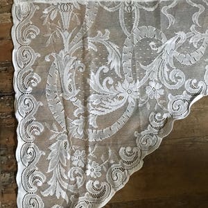 May include: White lace curtain with a floral pattern. The lace is delicate and intricate, with a scalloped edge.