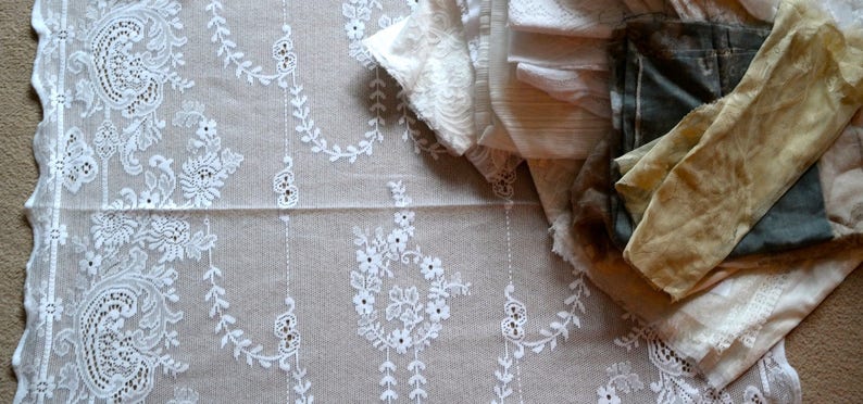 May include: A white lace tablecloth with a delicate floral pattern. A pile of white and beige fabric scraps are on top of the tablecloth.