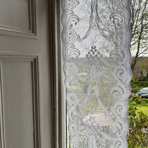 May include: White lace curtain with a delicate floral pattern hanging in a window. The curtain is sheer and allows light to pass through.