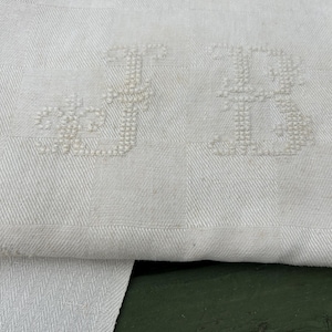 May include: Close-up of a white linen textile with a checkered pattern. The textile features embroidered initials "JB" in a delicate, ornate font. The fabric is folded, revealing a contrasting herringbone weave.