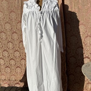 May include: A long, white nightdress with long sleeves and a frilled collar and trim. The nightdress is displayed on a mannequin. The fabric appears to be cotton or a similar lightweight material. The nightdress has a button-up front.