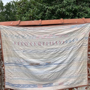 Pode incluir: Uma colcha têxtil retangular com uma base de cor creme e faixas horizontais de padrões costurados em tons suaves de azul, vermelho e castanho. O tecido apresenta pontos de acolchoado lineares visíveis e uma borda de tecido castanho sólido.