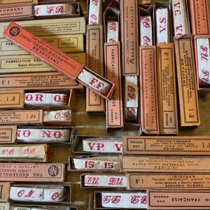 May include: A collection of vintage French fabric marking tapes in their original boxes. The boxes are in various shades of tan, peach, and red, with red text and lettering. The tapes are white with red letters and numbers. The boxes are labeled with French text.