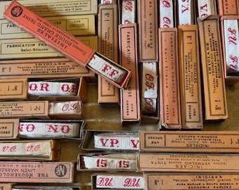 May include: A collection of vintage French fabric marking tapes in their original boxes. The boxes are in various shades of tan, peach, and red, with red text and lettering. The tapes are white with red letters and numbers. The boxes are labeled with French text.