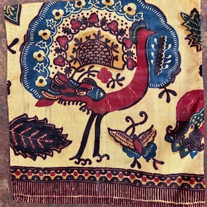 Puede incluir: Textil cuadrado estampado con un diseño de pájaro. El pájaro es rojo con patas negras y un elemento decorativo azul y amarillo detrás. El fondo es beige con detalles florales y mariposas azules, rojas y amarillas. Un borde rojo oscuro enmarca la parte inferior.