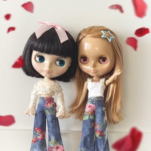 May include: Two fashion dolls with large eyes and unique features. One doll has black hair and a pink bow, the other has long blonde hair and a star hair clip. Both dolls wear denim pants with a floral pattern.