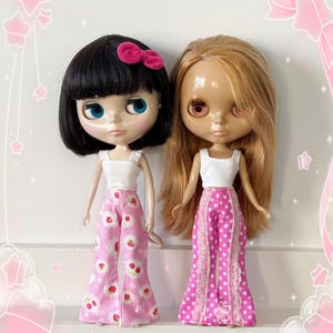 May include: Two Blythe dolls are displayed. One doll has black hair and a pink bow, the other has blonde hair. Both dolls wear white tops and pink pants. One pair of pants has a strawberry pattern, the other has white polka dots and lace.