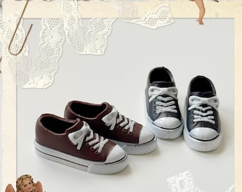 May include: Miniature brown and black sneakers with white laces and soles. The shoes are arranged on a white surface, with decorative elements including lace and angel figurines. The sneakers are a charming, small-scale representation of classic footwear.