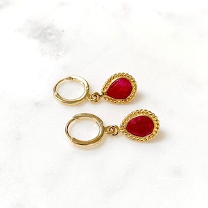 May include: A pair of gold-colored hoop earrings with teardrop-shaped red gemstones. The gemstones are framed by a twisted gold rope design. The earrings are displayed on a white marble surface.