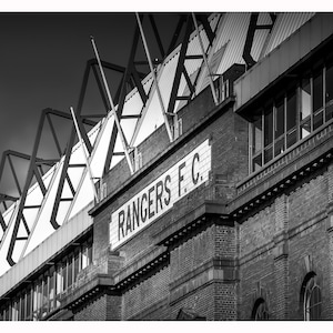 May include: Black and white photo of a brick building with a sign that reads "RANGERS F.C.". The building has a large, modern roof structure with a series of triangular supports.