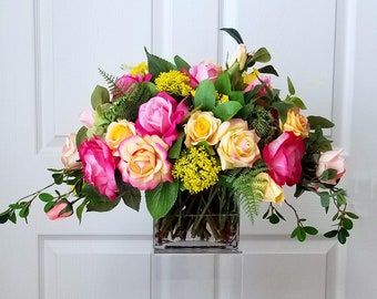 May include: A vibrant floral arrangement in a clear square glass vase. The bouquet features a mix of pink, yellow, and cream-colored roses, accented with green foliage and small yellow flowers. The arrangement is set against a white background.