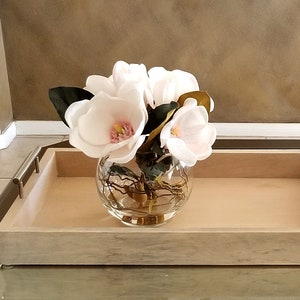 Puede incluir: Un cuenco de vidrio transparente lleno de flores de magnolia blancas con centros rosas y hojas verdes. El cuenco est&aacute; sobre una bandeja de madera clara con asas de metal, creando un arreglo floral decorativo.