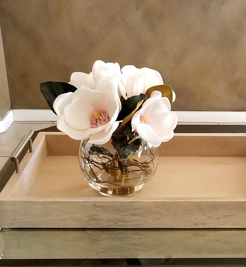 Puede incluir: Un cuenco de vidrio transparente lleno de flores de magnolia blancas con centros rosas y hojas verdes. El cuenco est&aacute; sobre una bandeja de madera clara con asas de metal, creando un arreglo floral decorativo.