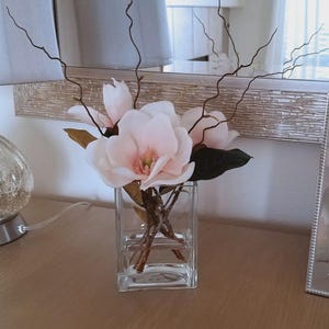 Puede incluir: Un jarr&oacute;n cuadrado de vidrio transparente contiene flores de magnolia rosas y ramitas marrones oscuras. El jarr&oacute;n est&aacute; sobre una superficie de madera, con un espejo plateado y una l&aacute;mpara decorativa en el fondo.
