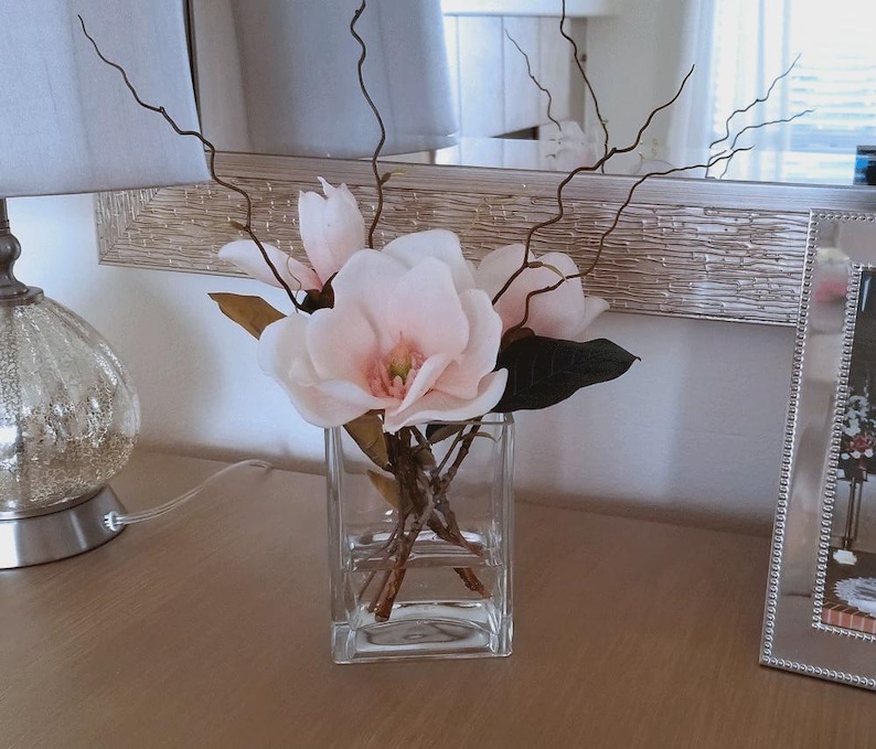 Puede incluir: Un jarr&oacute;n cuadrado de vidrio transparente contiene flores de magnolia rosas y ramitas marrones oscuras. El jarr&oacute;n est&aacute; sobre una superficie de madera, con un espejo plateado y una l&aacute;mpara decorativa en el fondo.
