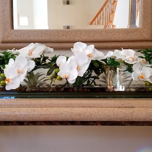 May include: A rectangular glass vase filled with white orchids, greenery, and small stones. The arrangement sits on a decorative table, with a mirror in the background. The orchids have yellow centers.