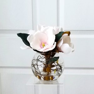 Puede incluir: Un jarr&oacute;n de vidrio transparente y redondo que contiene varias flores de magnolia blancas y rosas con hojas verdes. El jarr&oacute;n contiene ramitas decorativas y se encuentra sobre un soporte rectangular transparente. El fondo es una puerta blanca.