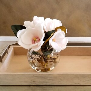 Puede incluir: Un cuenco de vidrio transparente lleno de flores de magnolia blancas con centros rosas y hojas verdes. El cuenco se encuentra sobre una bandeja de madera clara con asas de metal, creando un arreglo floral simple y elegante.