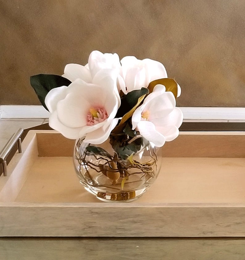 Puede incluir: Un cuenco de vidrio transparente lleno de flores de magnolia blancas con centros rosas y hojas verdes. El cuenco se encuentra sobre una bandeja de madera clara con asas de metal, creando un arreglo floral simple y elegante.