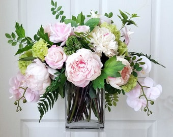 May include: A pink and white floral arrangement in a clear glass vase. The arrangement includes peonies, roses, orchids, and greenery.