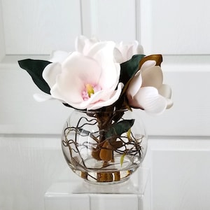 Puede incluir: Un arreglo floral en un cuenco de vidrio transparente con flores de magnolia artificiales. Las flores son blancas con centros rosas y amarillos. Tambi&eacute;n se ven hojas verdes oscuras y tallos marrones. El cuenco est&aacute; sobre un soporte cuadrado transparente.