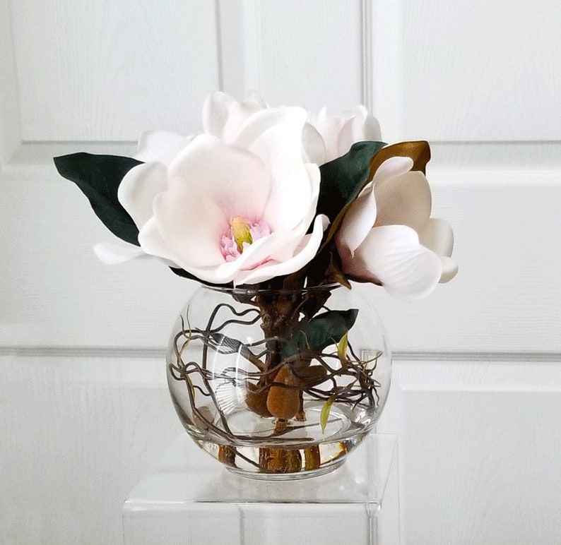 Puede incluir: Un arreglo floral en un cuenco de vidrio transparente con flores de magnolia artificiales. Las flores son blancas con centros rosas y amarillos. Tambi&eacute;n se ven hojas verdes oscuras y tallos marrones. El cuenco est&aacute; sobre un soporte cuadrado transparente.