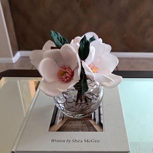 Puede incluir: Un jarr&oacute;n de cristal transparente lleno de flores de magnolia artificiales, con grandes p&eacute;talos de color rosa suave y hojas verdes. El jarr&oacute;n est&aacute; sobre un libro blanco con el texto "Written by Shea McGee". El arreglo est&aacute; sobre una mesa con tapa de cristal.