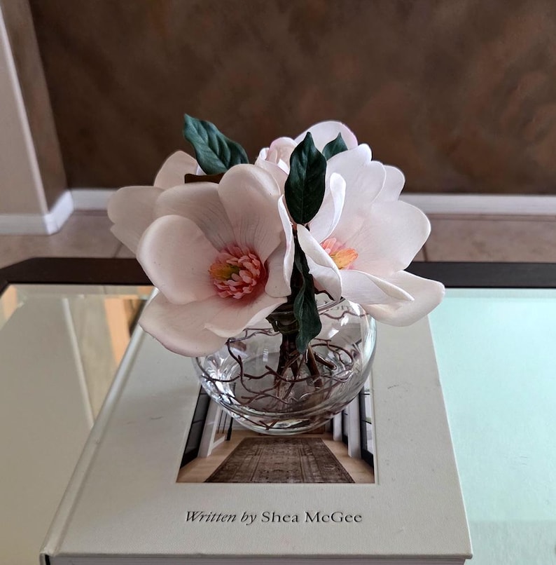 Puede incluir: Un jarr&oacute;n de cristal transparente lleno de flores de magnolia artificiales, con grandes p&eacute;talos de color rosa suave y hojas verdes. El jarr&oacute;n est&aacute; sobre un libro blanco con el texto "Written by Shea McGee". El arreglo est&aacute; sobre una mesa con tapa de cristal.