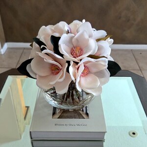 Puede incluir: Un cuenco de vidrio transparente lleno de flores de magnolia rosa claro con hojas de color verde oscuro. Las flores est&aacute;n sobre un libro con el texto "Written by Shea McGee" en una mesa con espejo.
