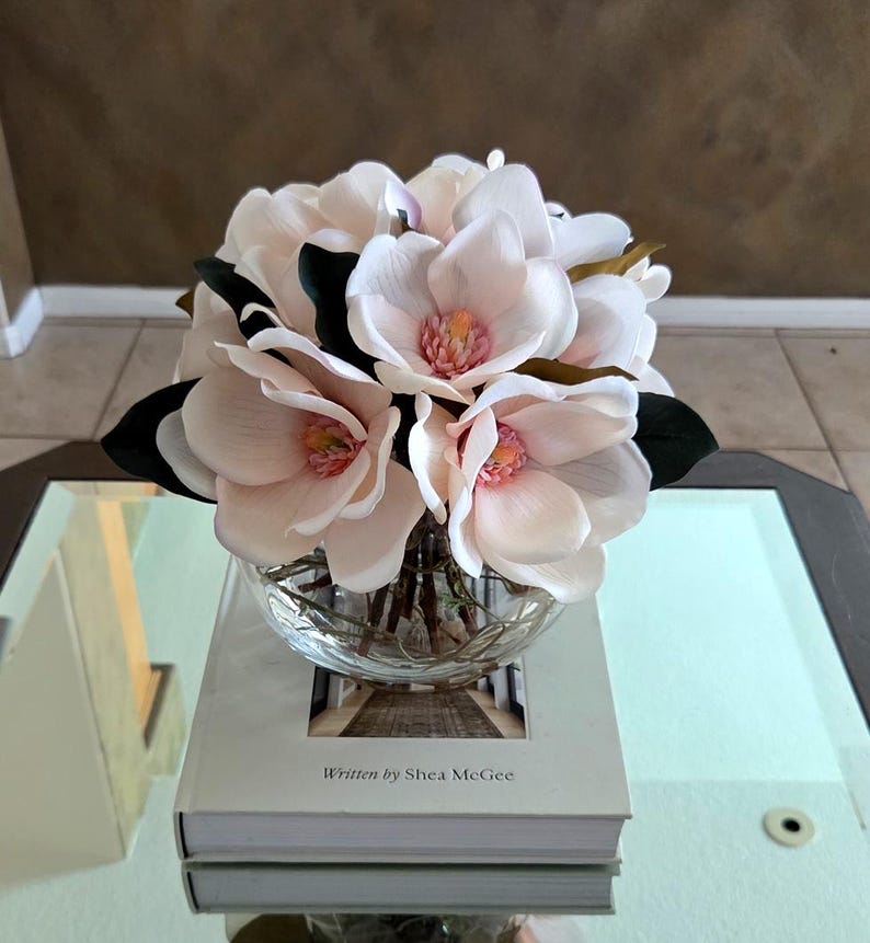 Puede incluir: Un cuenco de vidrio transparente lleno de flores de magnolia rosa claro con hojas de color verde oscuro. Las flores est&aacute;n sobre un libro con el texto "Written by Shea McGee" en una mesa con espejo.