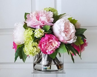 May include: A bouquet of pink and white peonies and green hydrangeas in a clear glass vase. The flowers are arranged in a circular shape and are sitting on a white surface.