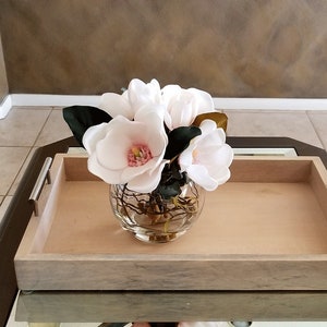 Puede incluir: Un arreglo floral con flores de magnolia blancas con centros rosas y hojas verde oscuro en un jarr&oacute;n de vidrio transparente y redondo. El jarr&oacute;n est&aacute; sobre una bandeja de madera clara con asas plateadas.