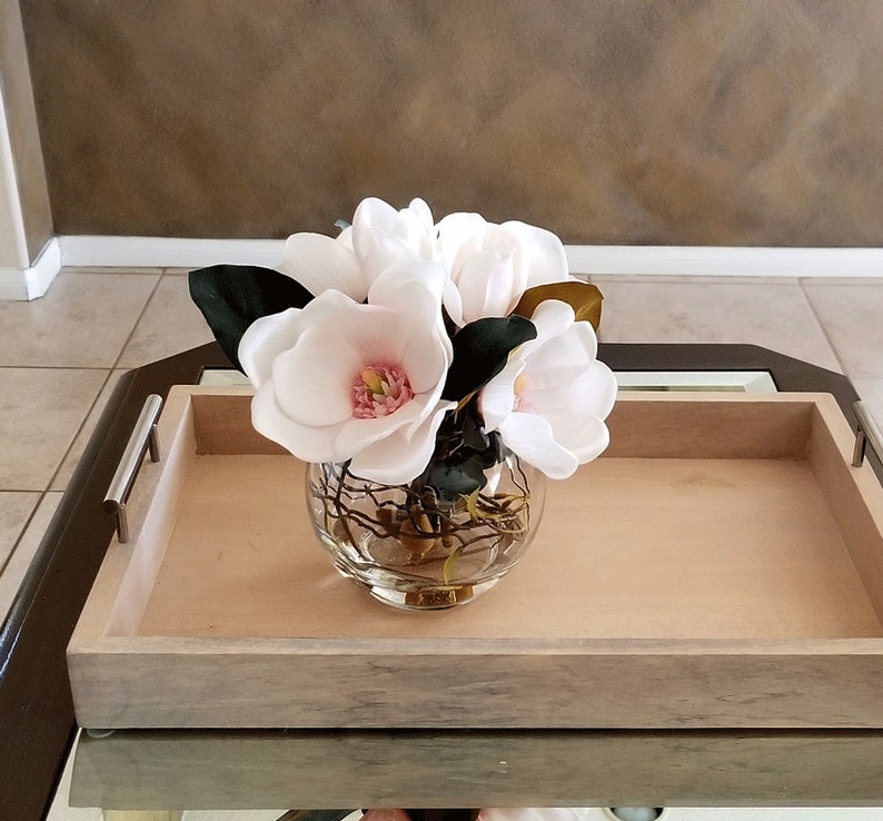 Puede incluir: Un arreglo floral con flores de magnolia blancas con centros rosas y hojas verde oscuro en un jarr&oacute;n de vidrio transparente y redondo. El jarr&oacute;n est&aacute; sobre una bandeja de madera clara con asas plateadas.