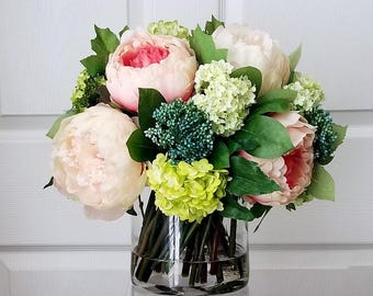 May include: A bouquet of pink and white peonies and green hydrangeas in a clear glass vase. The flowers are arranged in a symmetrical pattern and are surrounded by green leaves.
