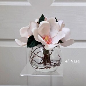 Puede incluir: Un jarr&oacute;n de vidrio transparente y redondo, de aproximadamente 10 cm de di&aacute;metro, contiene un ramo de flores de magnolia artificiales. Las flores son de color rosa p&aacute;lido con hojas verdes y un centro amarillo. Hay ramitas dentro.
