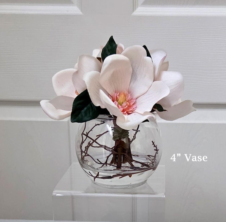 Puede incluir: Un jarr&oacute;n de vidrio transparente y redondo, de aproximadamente 10 cm de di&aacute;metro, contiene un ramo de flores de magnolia artificiales. Las flores son de color rosa p&aacute;lido con hojas verdes y un centro amarillo. Hay ramitas dentro.