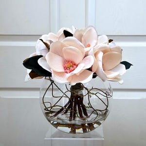 Puede incluir: Arreglo floral con grandes flores de magnolia rosa claro con hojas verdes oscuras y marrones. Las flores est&aacute;n dispuestas en un jarr&oacute;n de vidrio transparente y redondo con ramitas decorativas y agua, sobre un soporte acr&iacute;lico transparente.