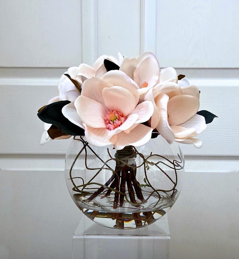 Puede incluir: Arreglo floral con grandes flores de magnolia rosa claro con hojas verdes oscuras y marrones. Las flores est&aacute;n dispuestas en un jarr&oacute;n de vidrio transparente y redondo con ramitas decorativas y agua, sobre un soporte acr&iacute;lico transparente.
