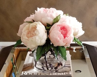 May include: A bouquet of pink and white peonies in a clear glass vase on a table with a magazine. The magazine cover reads "Room & Board".