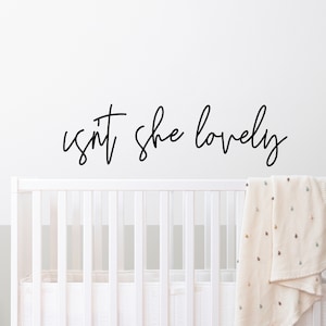 May include: A nursery wall decal with the phrase "isn't she lovely" in a cursive font. Below the text is a white crib with a blanket draped over the side. A wooden dresser and decorative cactus are in the background.