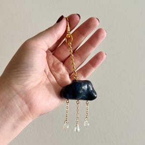 May include: A dark blue cloud-shaped keychain with gold-colored chain and clasp. The cloud has three dangling strands with clear, teardrop-shaped beads. The keychain is held up against a neutral background.