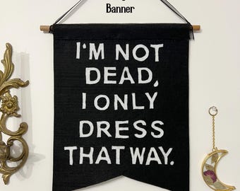 May include: Black felt banner with white text that reads "I'm not dead, I only dress that way." The banner is hanging on a white wall with a wooden dowel rod at the top.