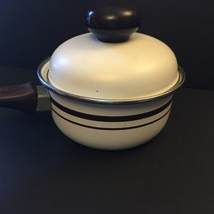 May include: A white saucepan with a lid and a dark brown handle. The pot has two dark brown stripes around the middle and a dark brown knob on the lid. The pot is sitting on a black surface.